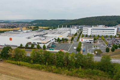 Fabrikgelände, das in einer Landschaft auf Felder und Wald liegt. Im Hintergrund ist ein Dorf.