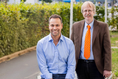 Daniel Fischer sitzt auf einer Bank. Alexander Bracht steht neben ihm. Dahinter ist eine Hecke.